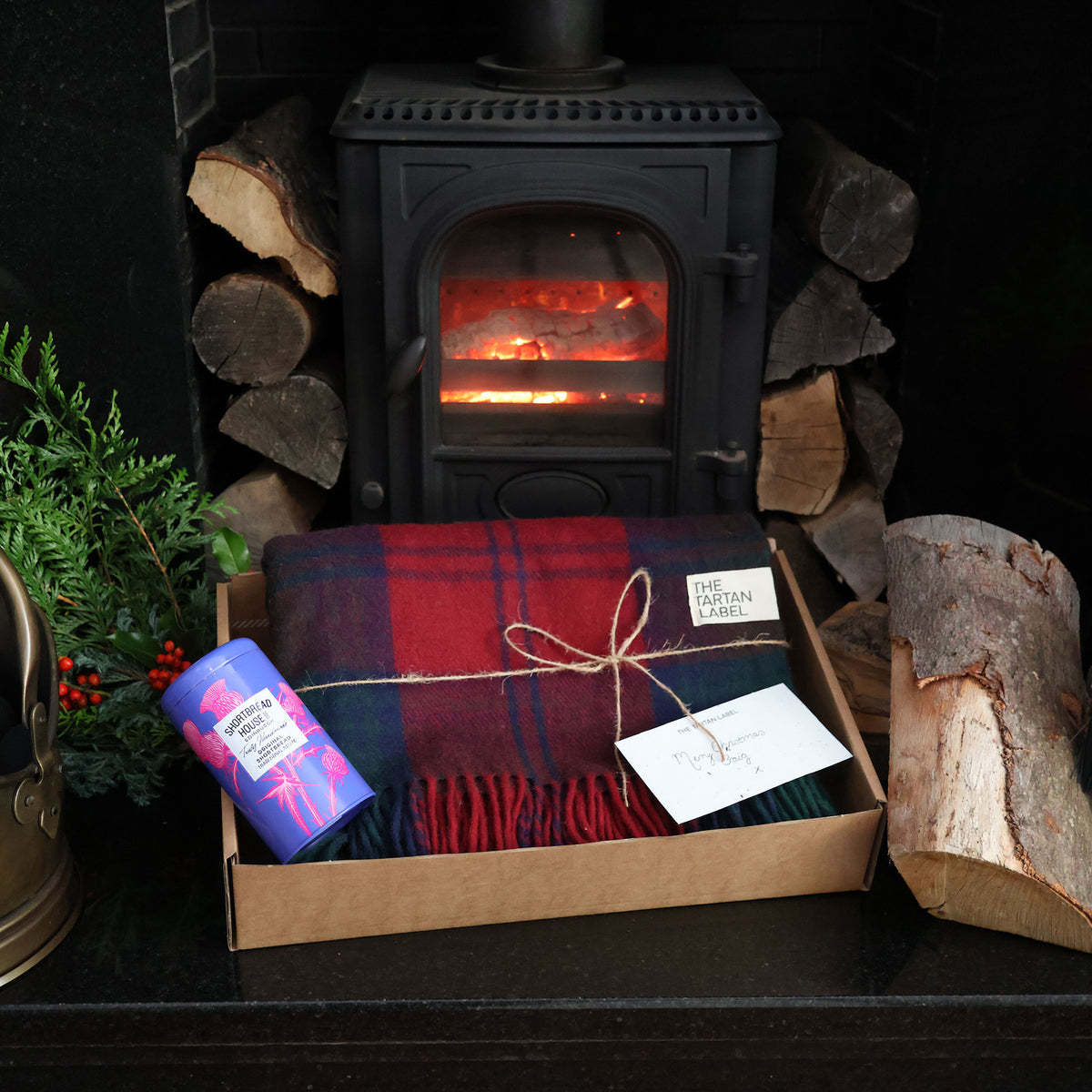 A cozy gift box set on a fireplace mantel, featuring a red and navy plaid tartan throw blanket wrapped in twine, a shortbread tin, and a card, set against a black wood stove with logs and greenery.