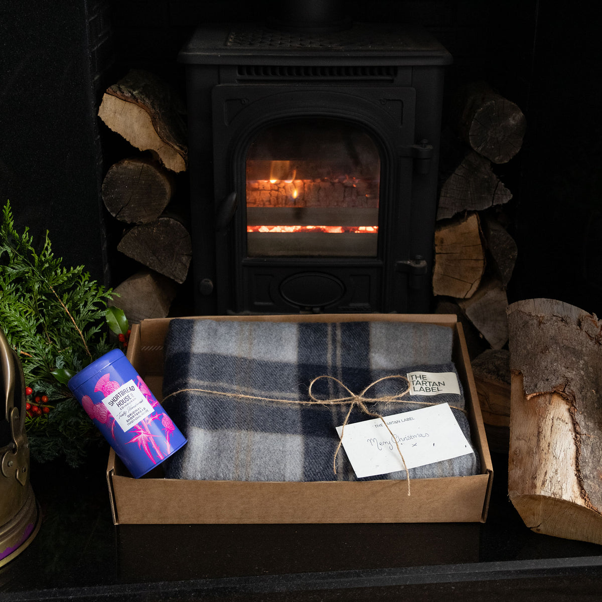 A cozy fireplace scene with a gift box containing a folded tartan blanket in blue, grey, and white tones, tied with twine and a small tag. A shortbread tin sits beside it, with stacked firewood and a black wood-burning stove in the background.