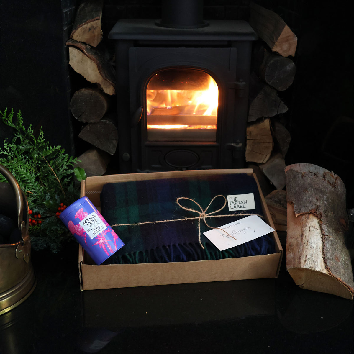A gift set featuring a tartan plaid throw blanket in a brown box, alongside a shortbread tin and a small card, styled in front of a burning wood-burning stove.